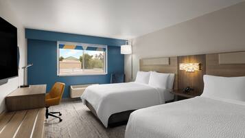 A hotel room with two beds, a desk, a chair, a TV, and a window with a view of trees and a building.