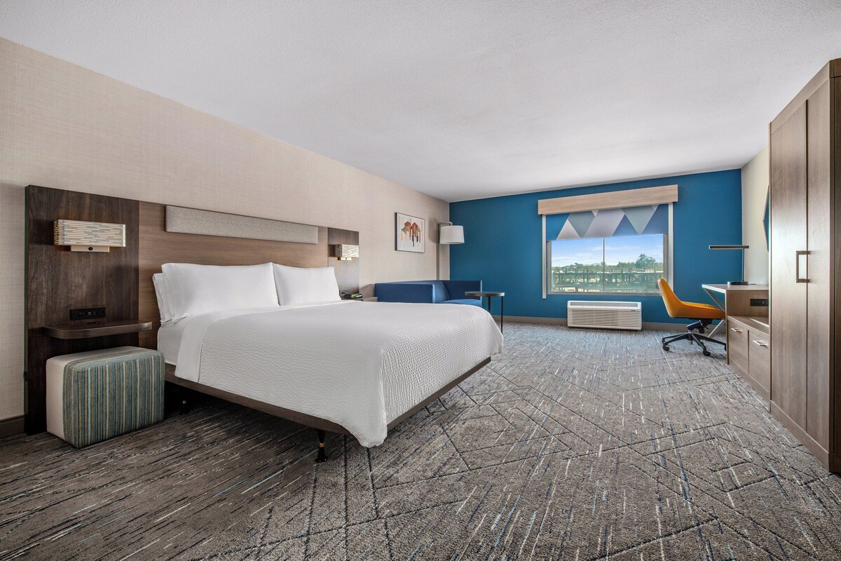 A hotel room with a large bed, a desk, a chair, a window with a view, and a small table with a lamp.