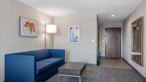 A modern hotel room with a blue sofa, a small table, and a lamp. There are two framed artworks on the wall and a wooden door in the background. - Holiday Inn Express Hotel & Suites San Diego-Sorrento Valley by IHG (San Diego)