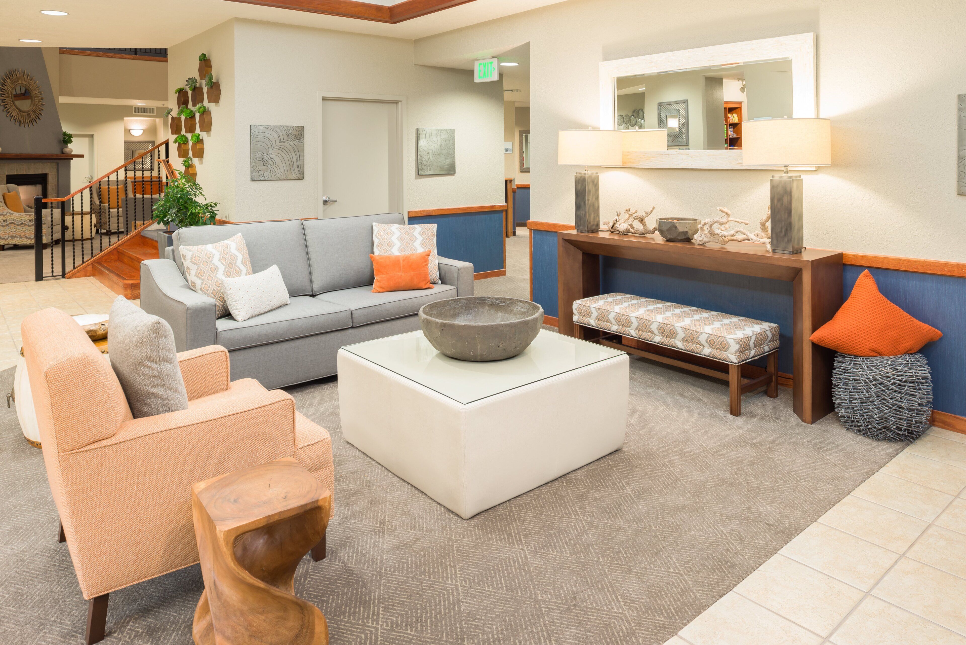 A modern hotel lobby with a grey sofa, a white ottoman, and a wooden coffee table. There is a staircase with a wooden railing leading to an upper level.
