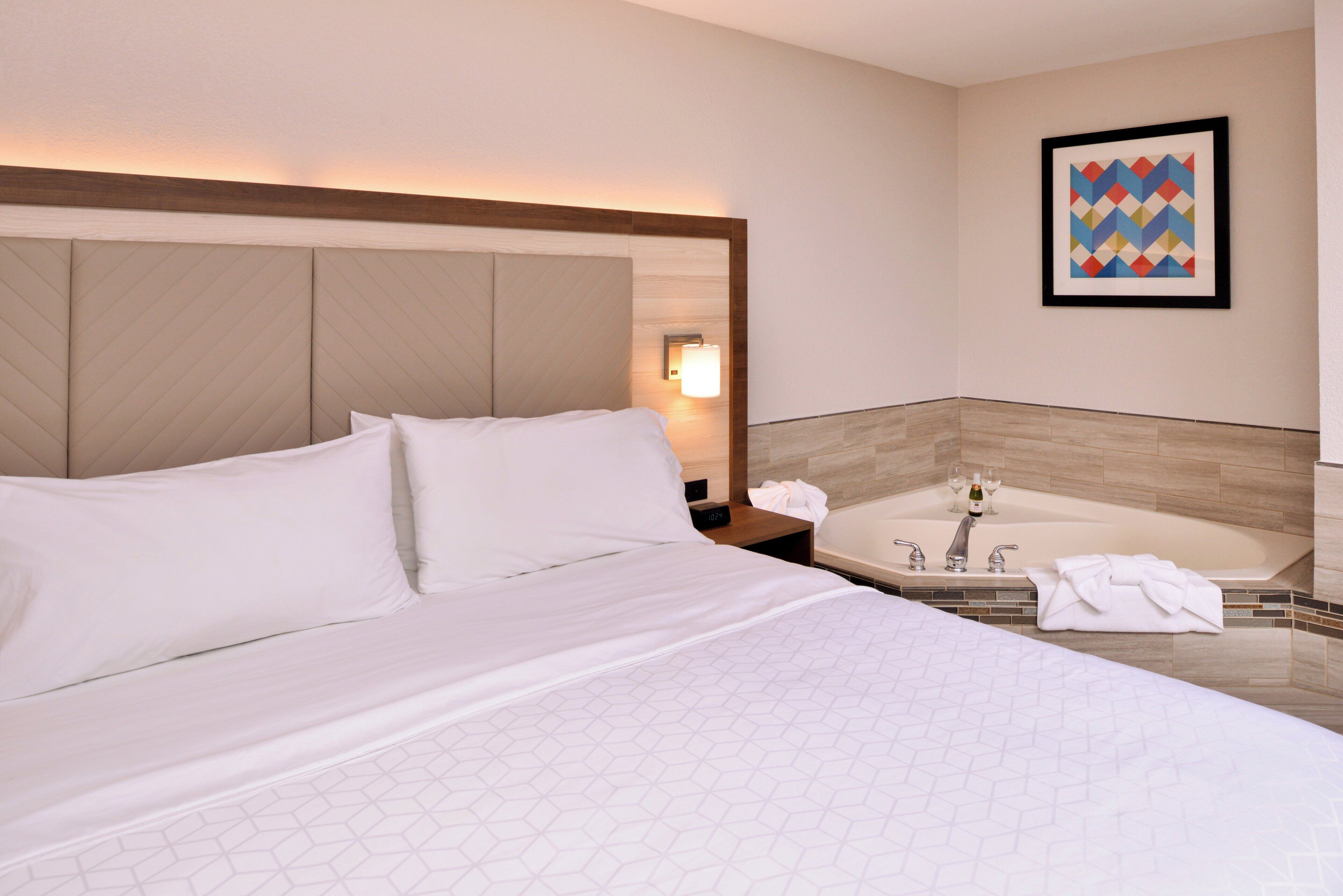 A hotel room with a large bed, a nightstand with a lamp, a framed picture on the wall, and a bathtub.