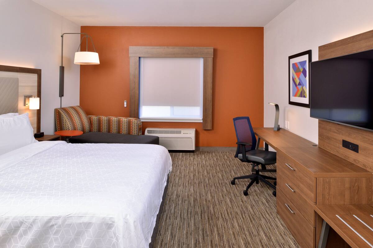 A hotel room with a large bed, a desk with a chair, a television, and a window with blinds.