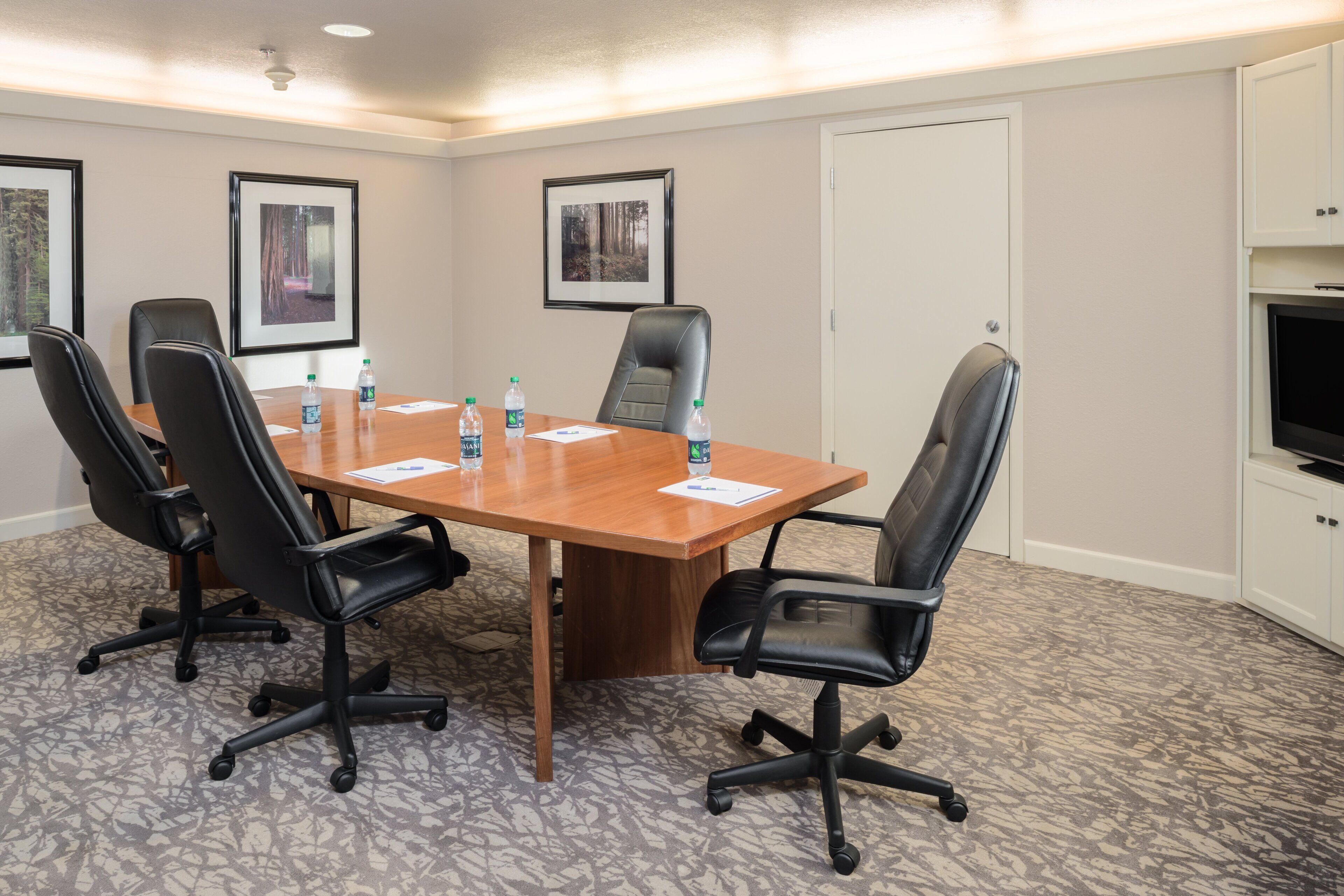 A conference room with a wooden table, black chairs, water bottles, framed pictures on the wall, and a television on the right.