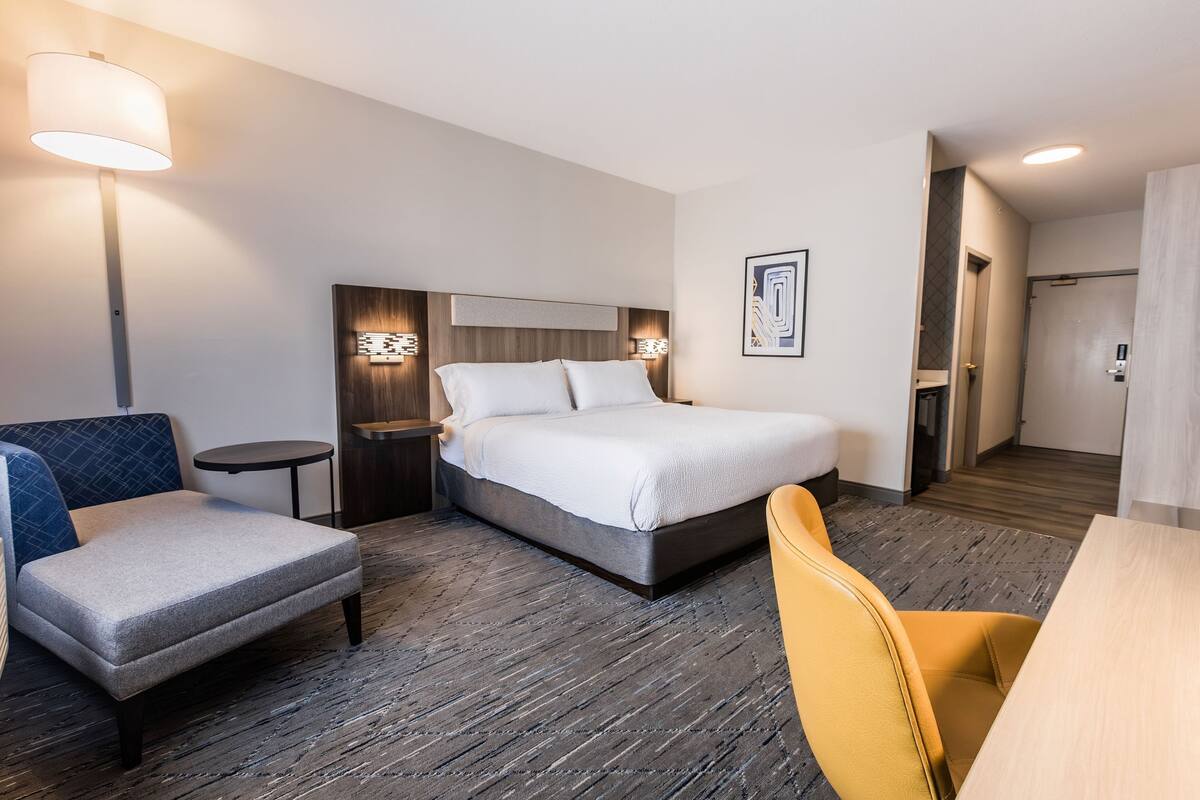 A hotel room with a bed, a chair, a small table, and a lamp.