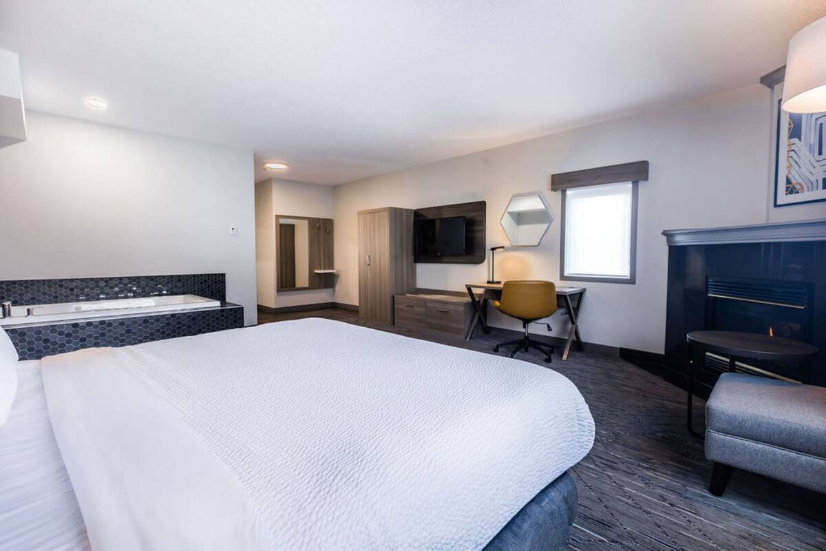 A hotel room with a large bed, a desk with a chair, a TV, a fireplace, and a bathroom with a bathtub.