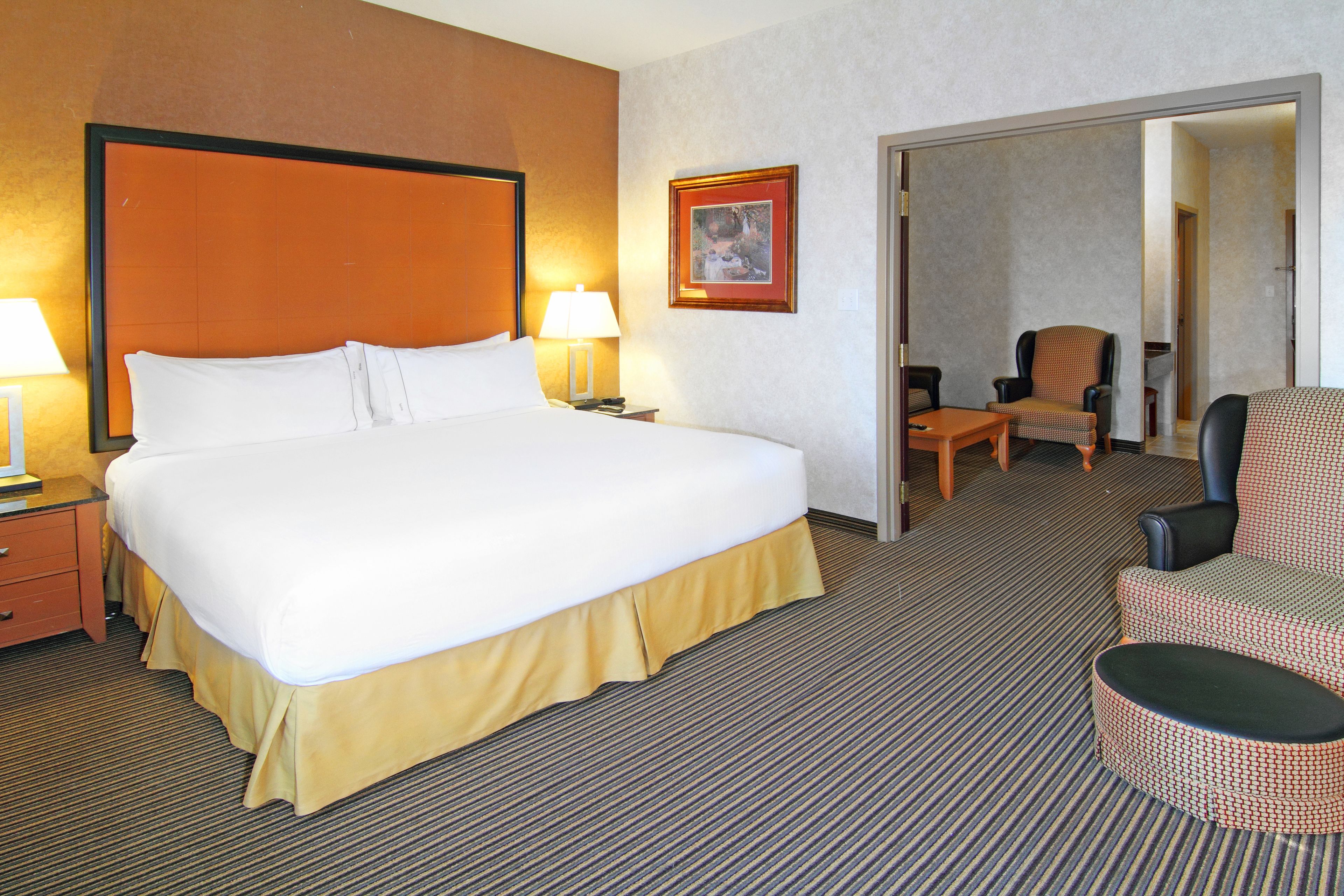 Habitación de hotel con una cama grande, dos mesitas de noche, una silla y una puerta que da a otra habitación.