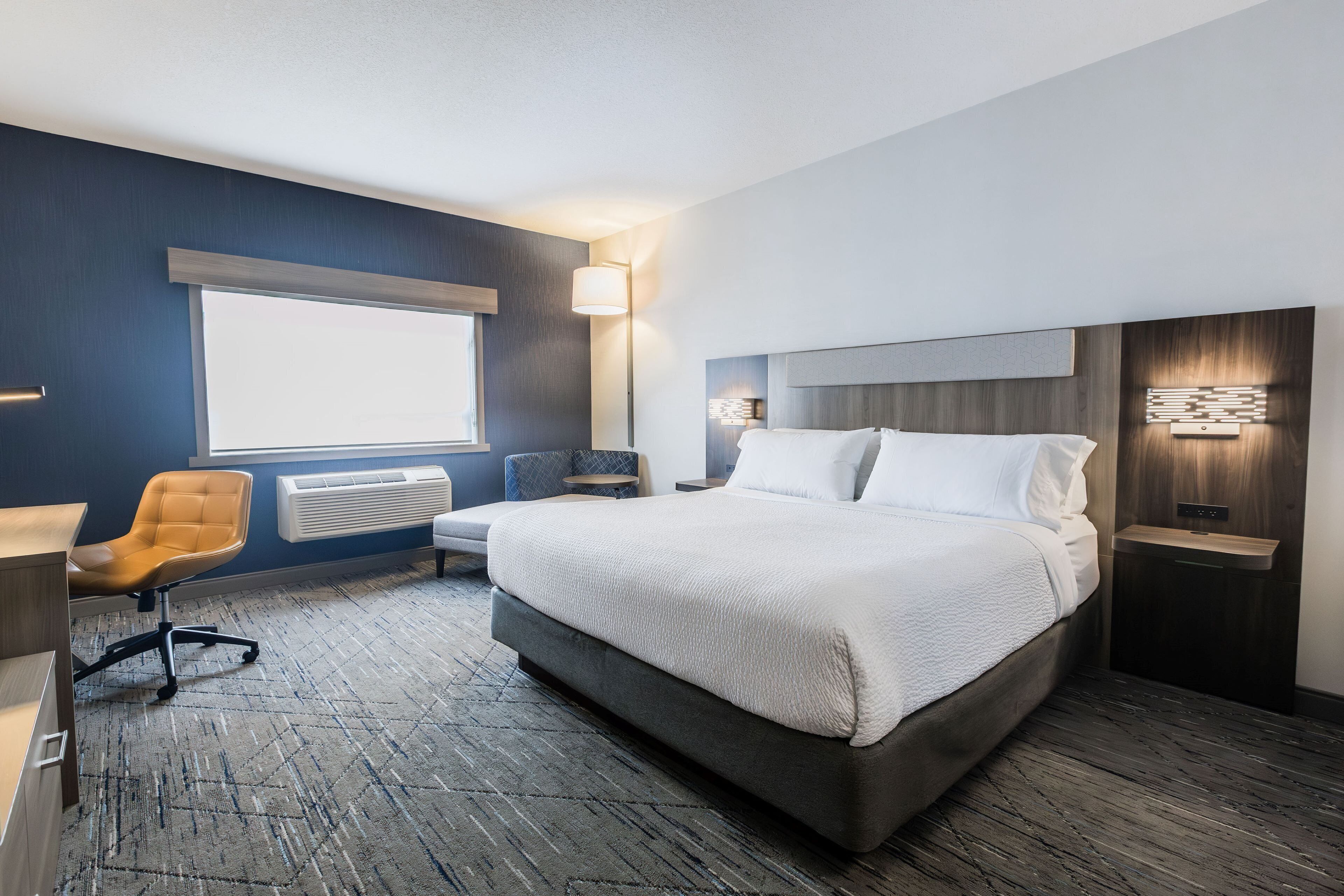 A modern hotel room with a large bed, a desk, a chair, and a window.
