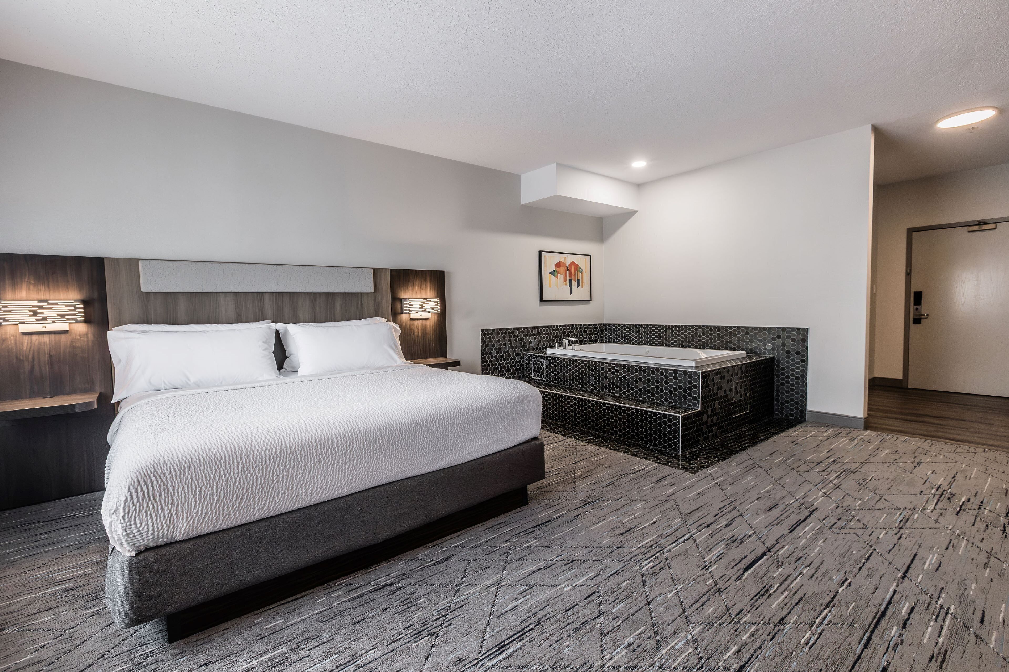 A modern hotel room with a large bed, a small bathroom with a bathtub, and a wooden floor.