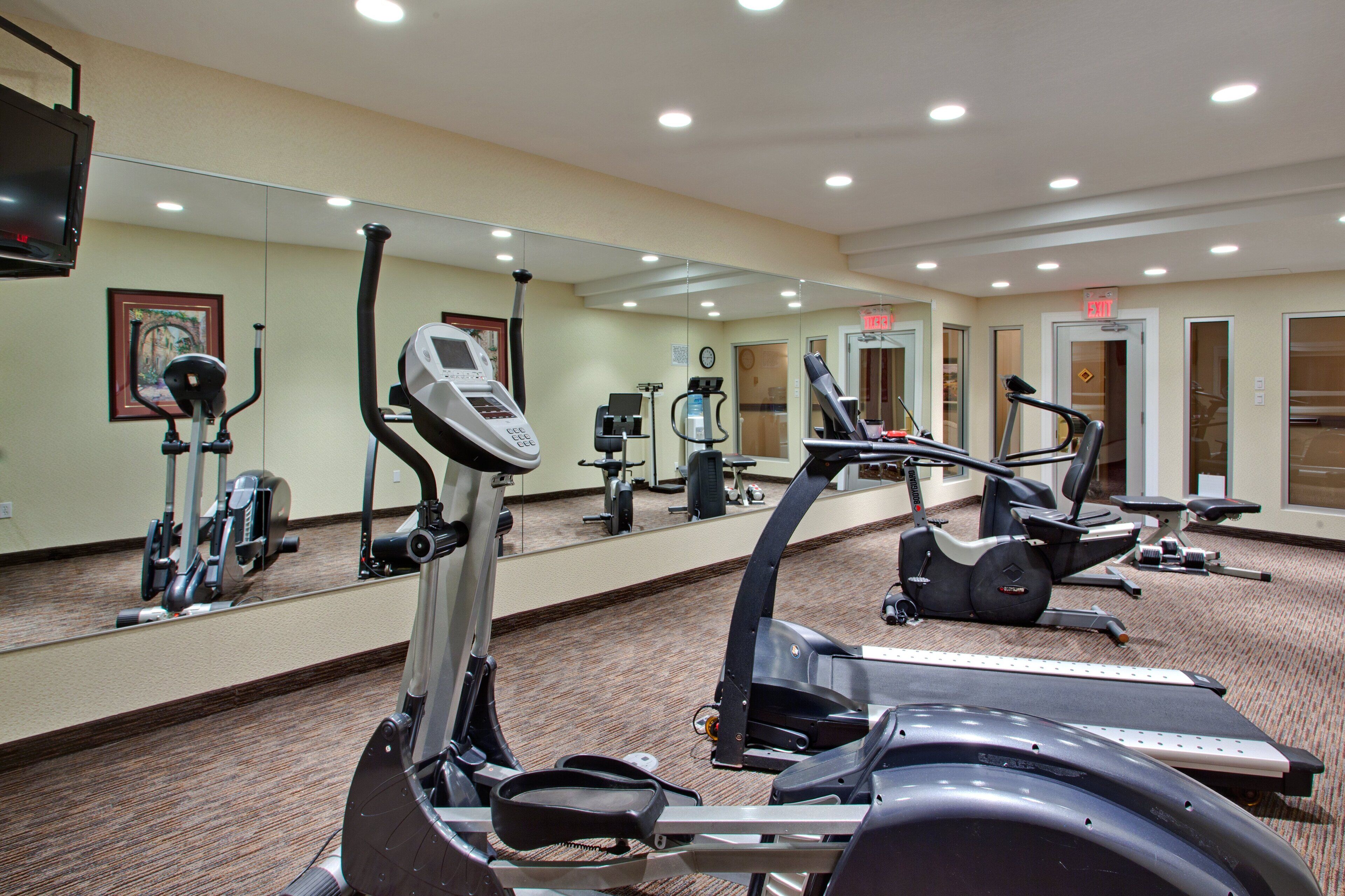 A hotel fitness center with multiple exercise machines, a large mirror, and an exit sign.