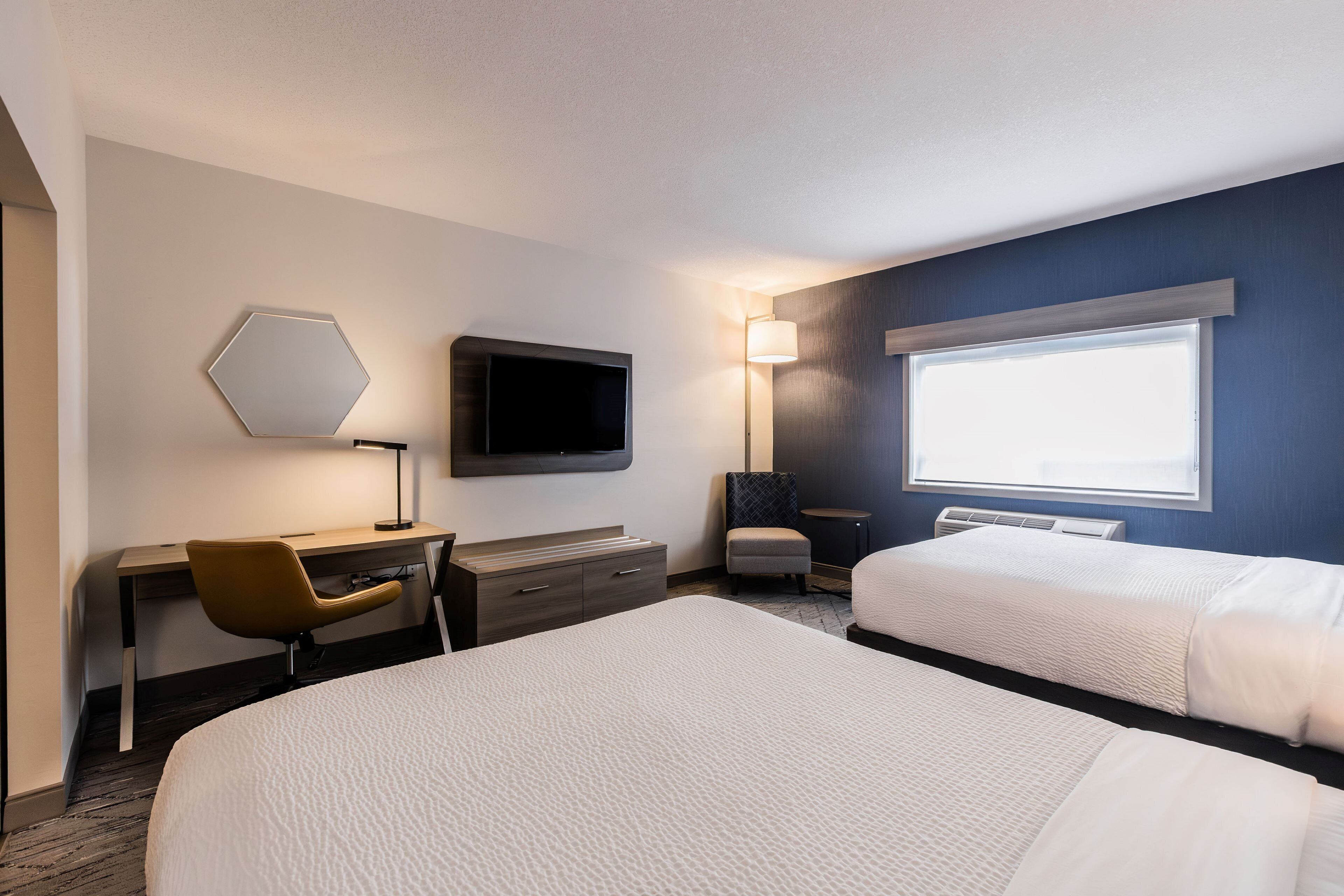 Habitación de hotel con cama, escritorio, silla, televisión, lámpara y ventana.