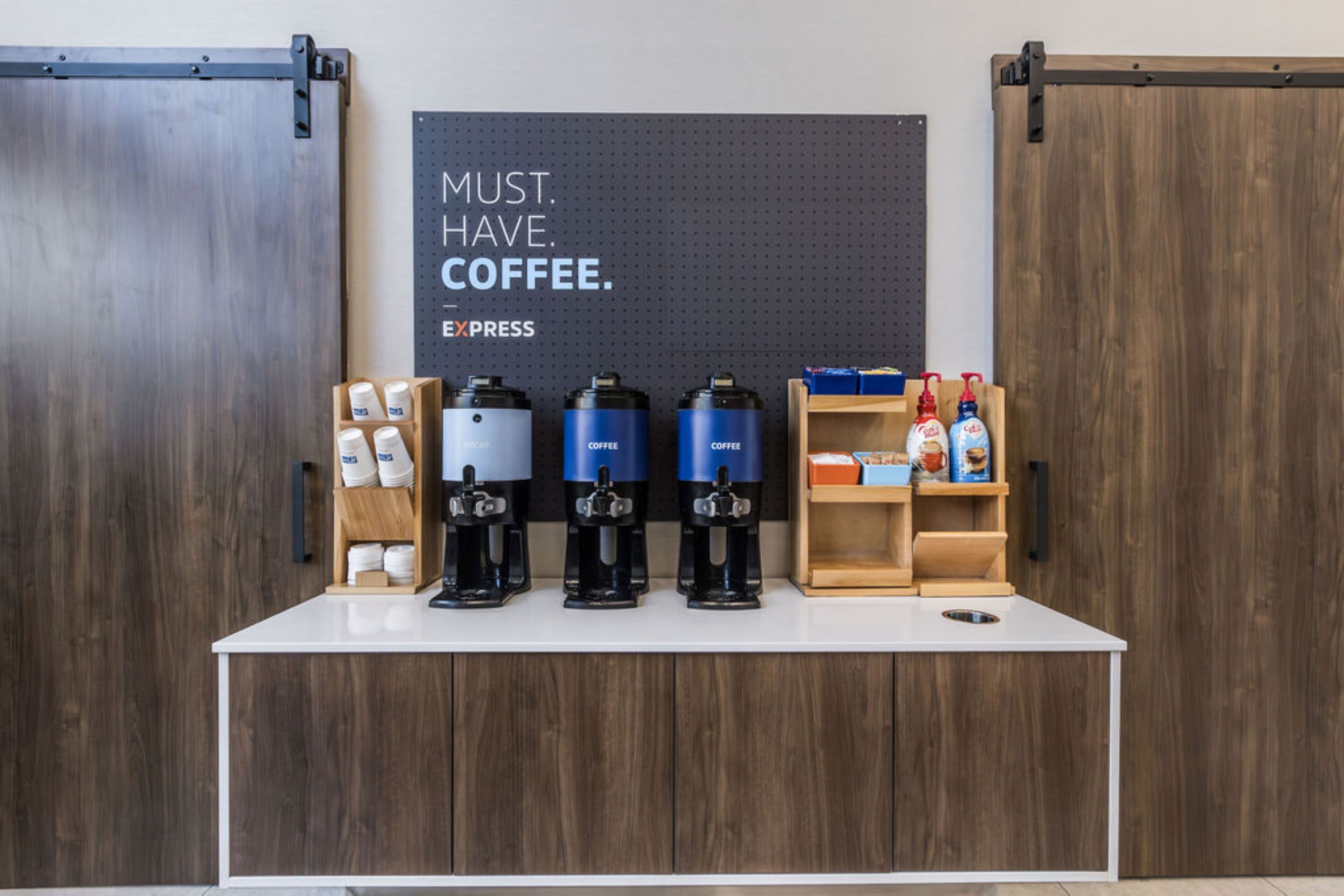 A coffee station with four coffee machines, a sign that says "MUST HAVE COFFEE," and a shelf with coffee pods and a bottle of hand sanitizer.