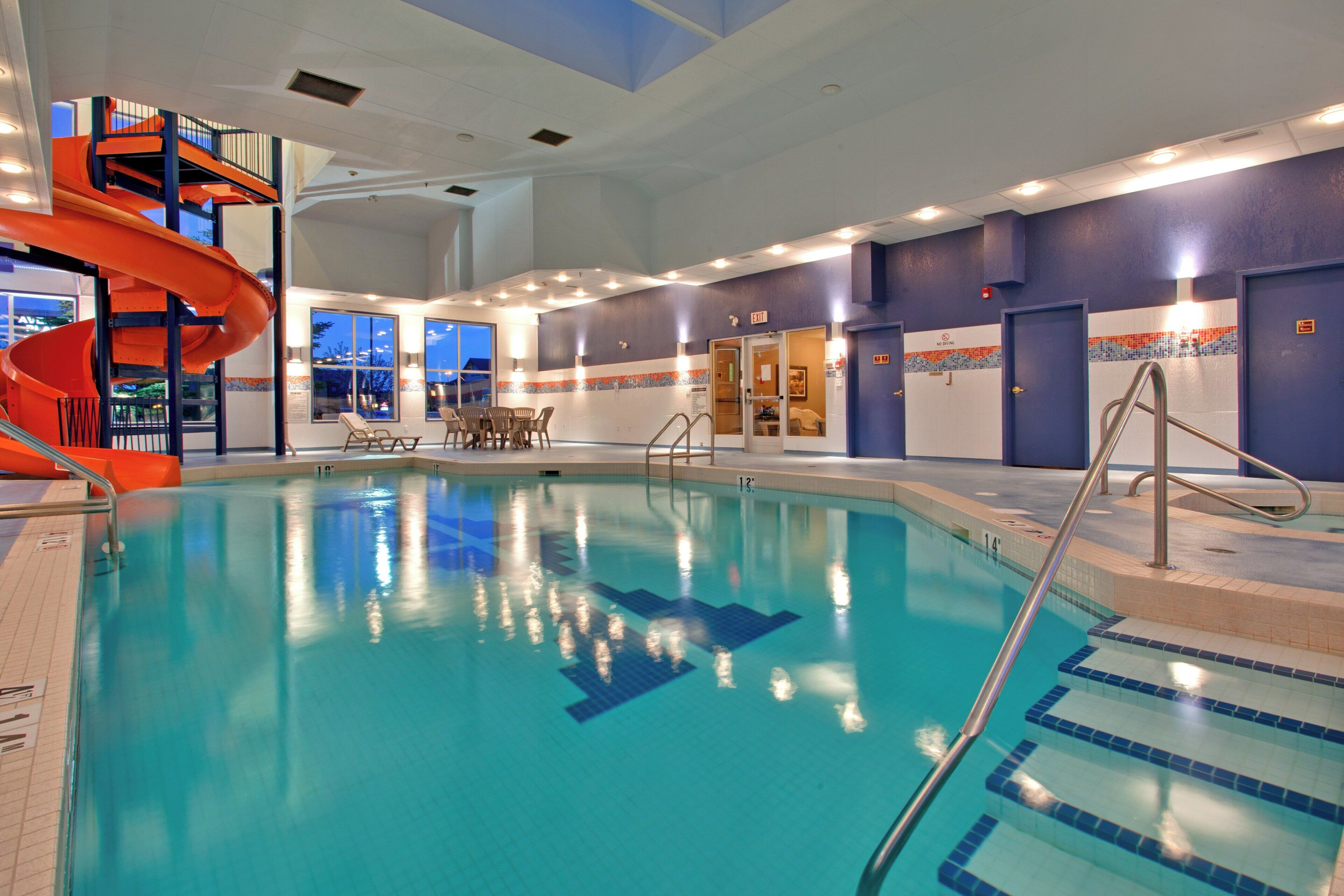 Una piscina interior con un tobogán en espiral, múltiples salidas y una zona de estar con mesas y sillas.