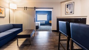 Una camera d'albergo con un divano blu, un tavolino in legno, una scrivania e una sedia. Si vede un letto con una testiera bianca e una finestra con persiane.