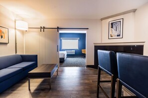 Een hotelkamer met een blauwe bank, een houten salontafel, een bureau en een stoel. Er is een bed met een wit hoofdeinde en een raam met zonwering.