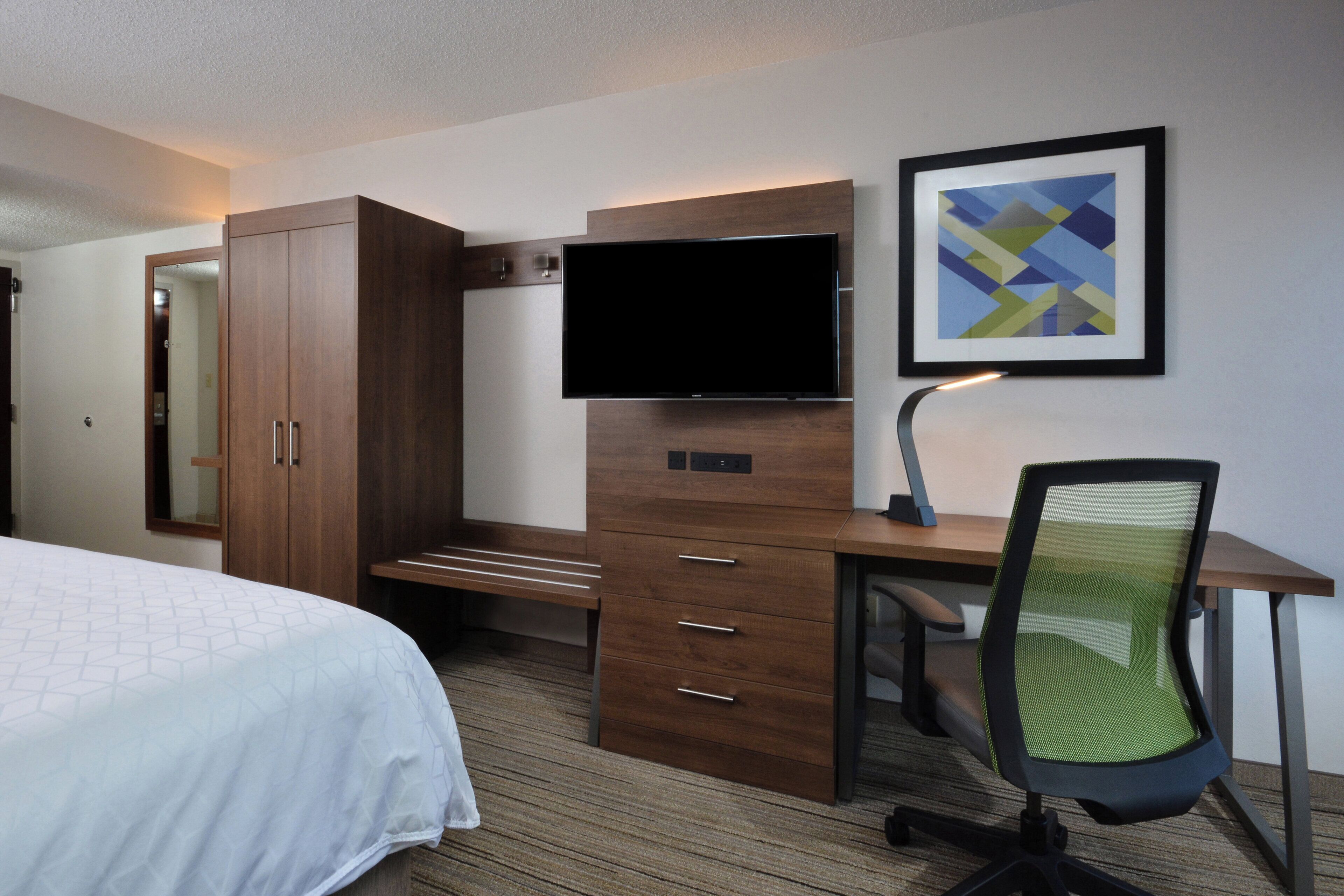 A hotel room with a bed, a desk with a chair, a television, and a framed picture on the wall.