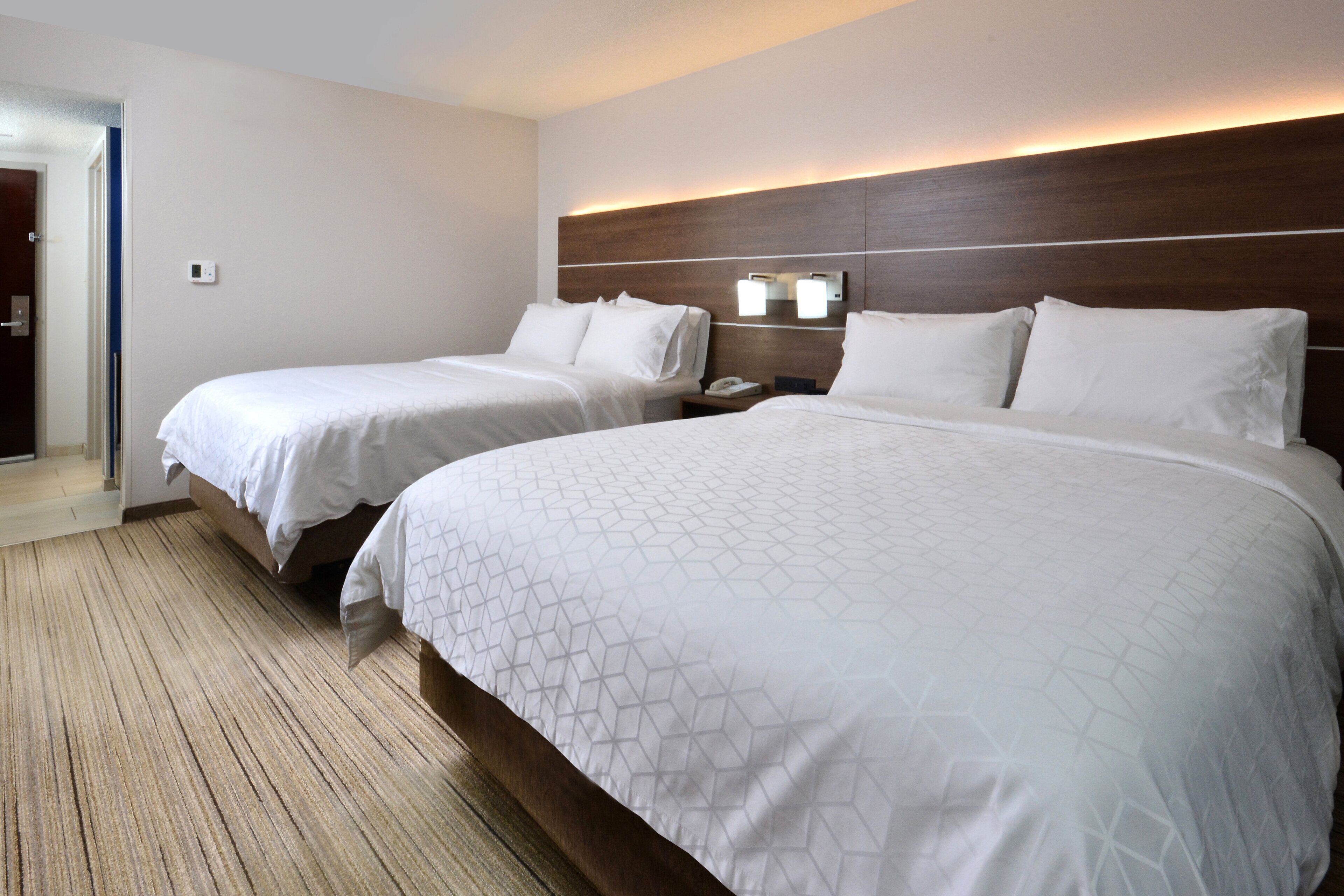 Quarto de hotel com duas camas, cabeceira de madeira, mesa de cabeceira com telefone e piso coberto por carpete.
