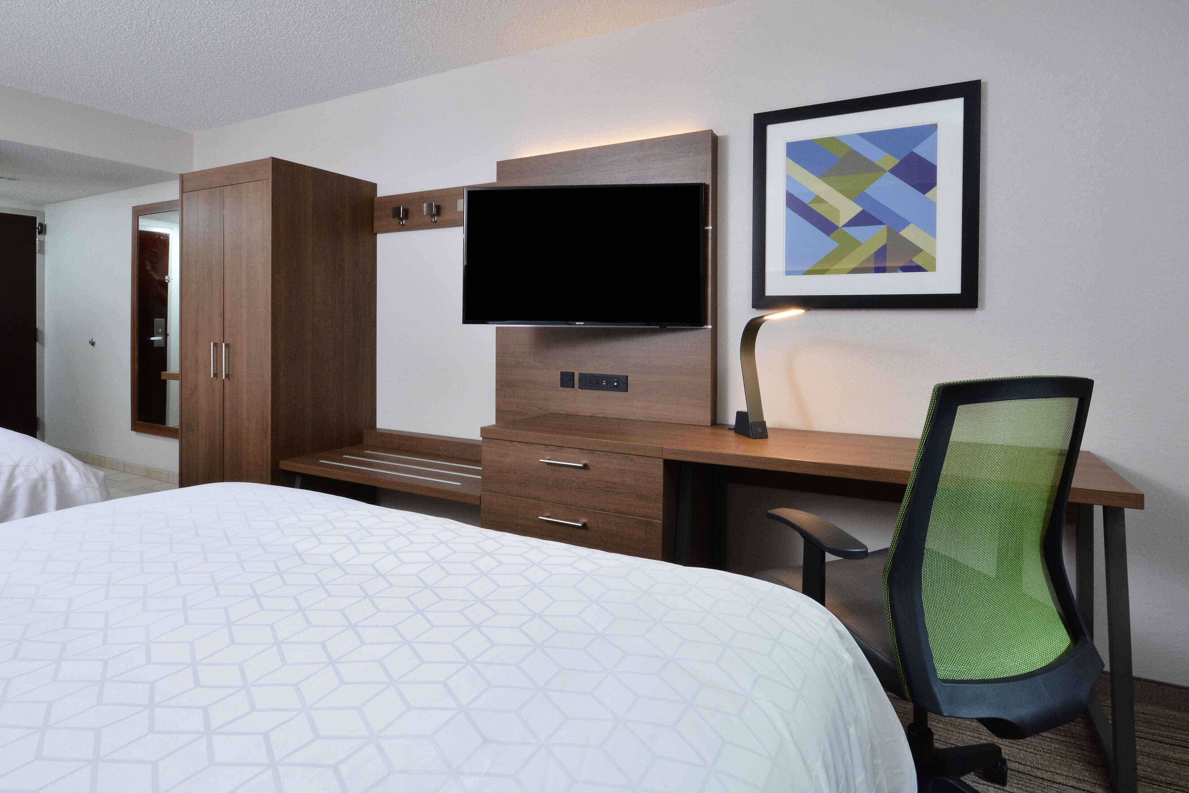 A hotel room with a bed, a desk with a flat-screen TV, a chair, and a framed picture on the wall.