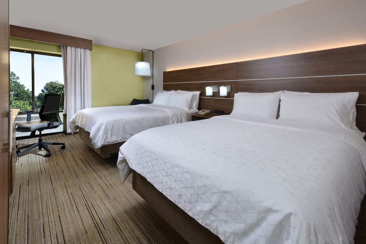 A hotel room with two beds, a desk with a chair, and a large window with curtains.