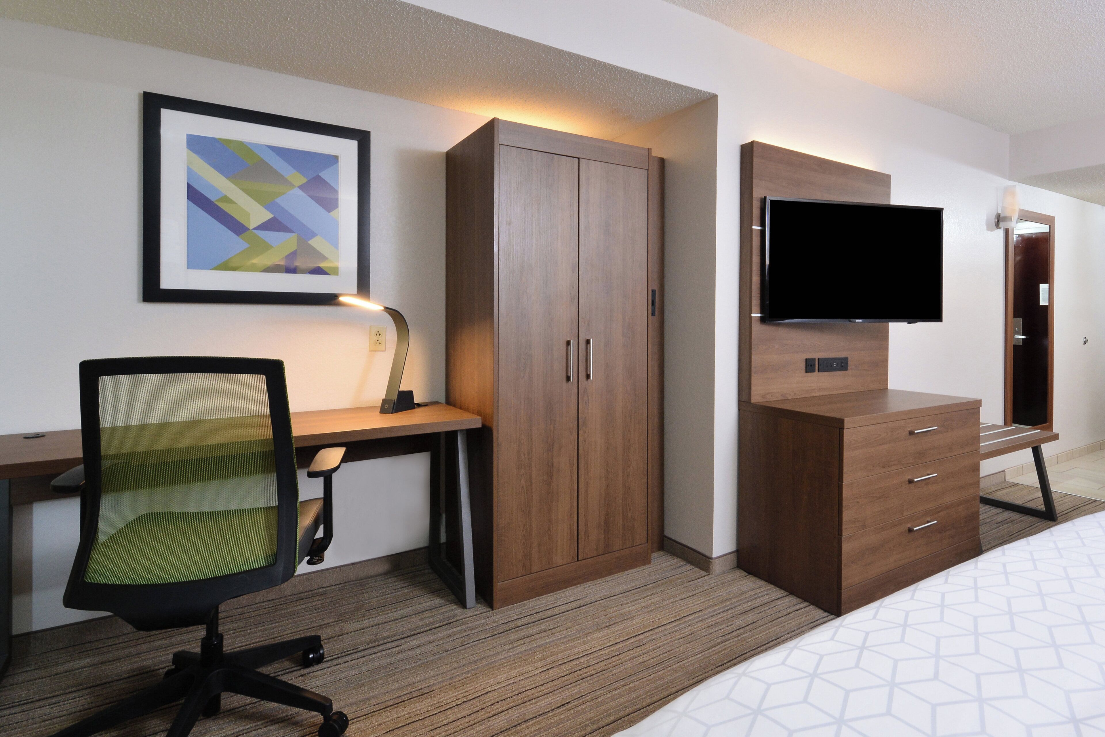 A hotel room with a bed, a desk with a chair, a wardrobe, a television, and a framed picture on the wall.