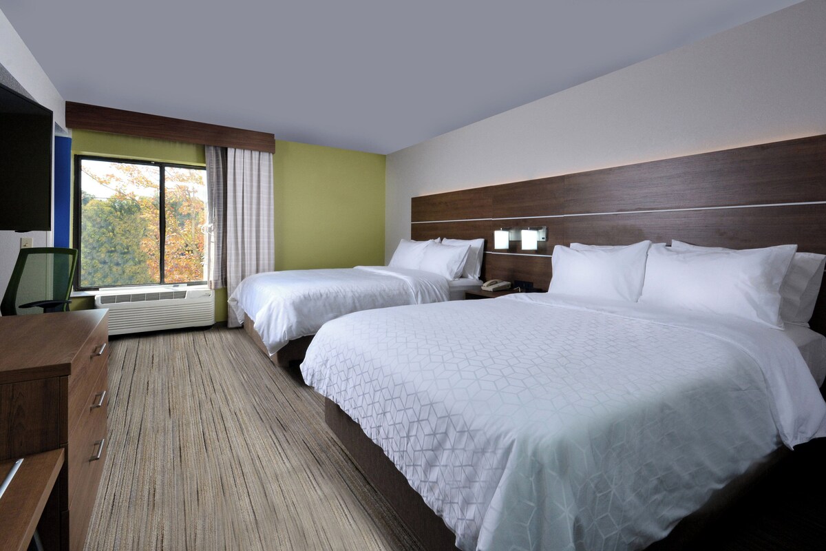 A hotel room with two beds, a desk, a TV, and a window with a view of trees.