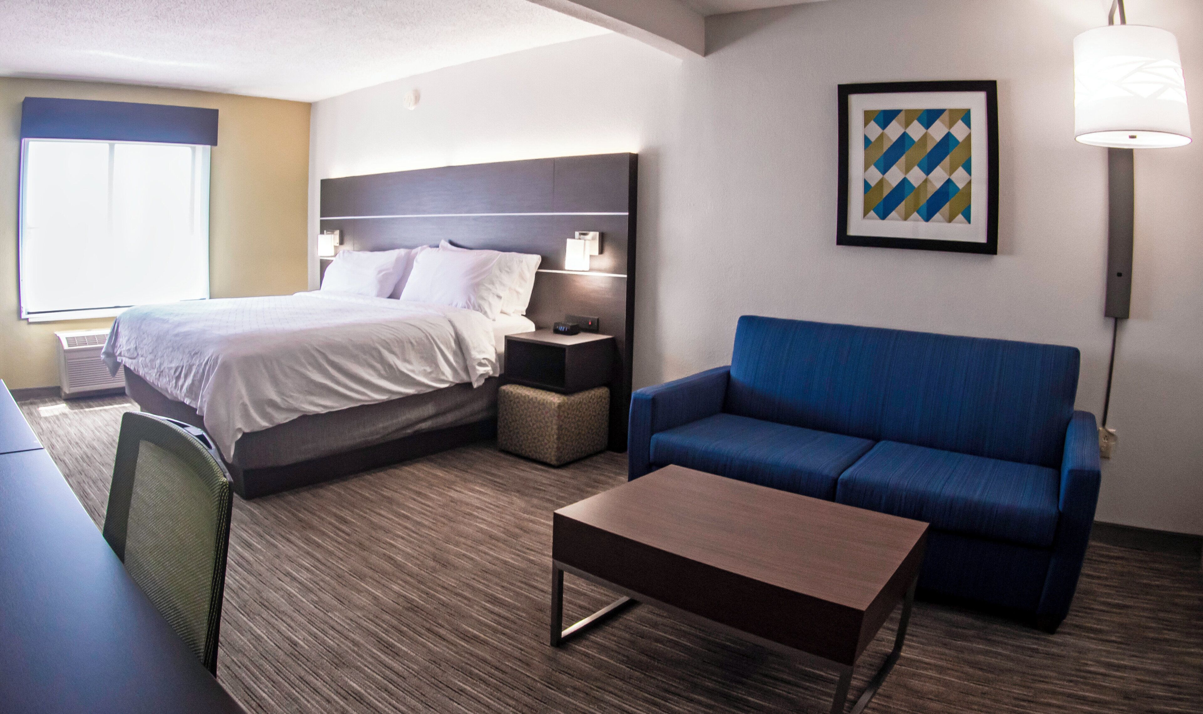 A hotel room with a bed, a blue sofa, a small table, a chair, and a picture on the wall.