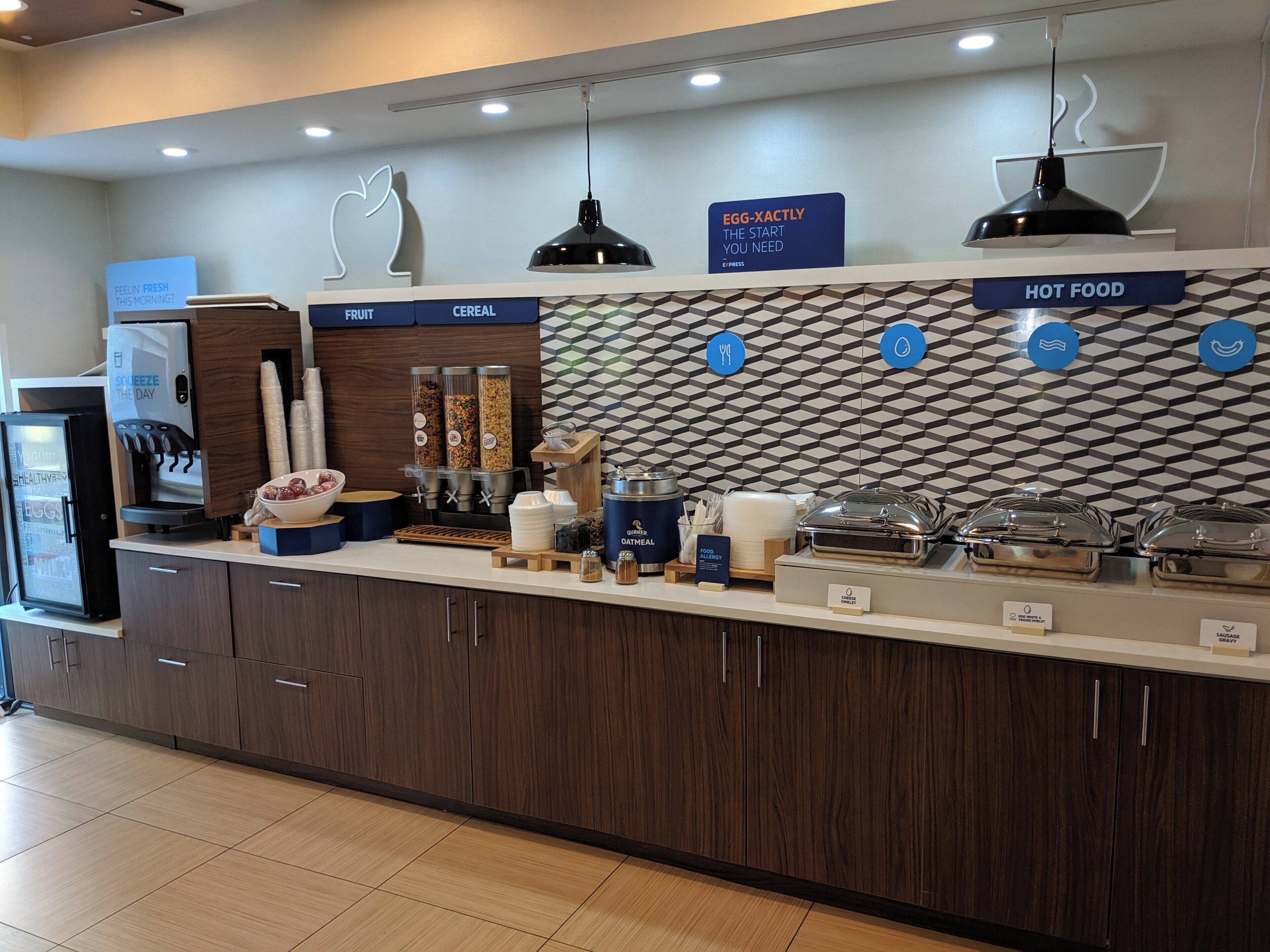 Una barra de desayuno con secciones identificadas para fruta, cereales y alimentos calientes, equipada con una máquina de café y dispensador de bebidas.