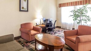 A hotel room with two pink armchairs, a wooden coffee table, a desk with a computer and printer, a lamp, and a large window with a view of a tree.