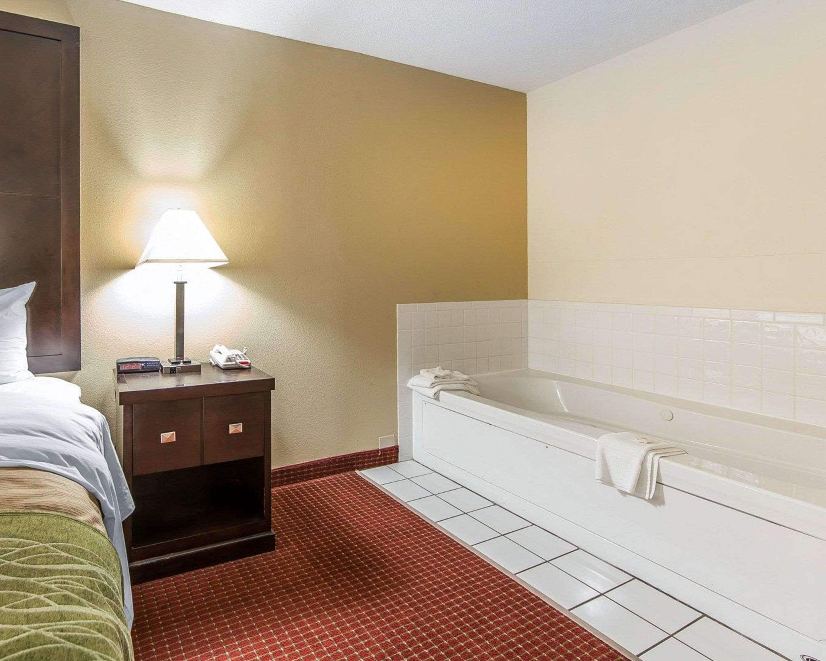 Une chambre d’hôtel avec un lit, une table de chevet avec une lampe, un téléphone et une baignoire blanche avec une serviette.
