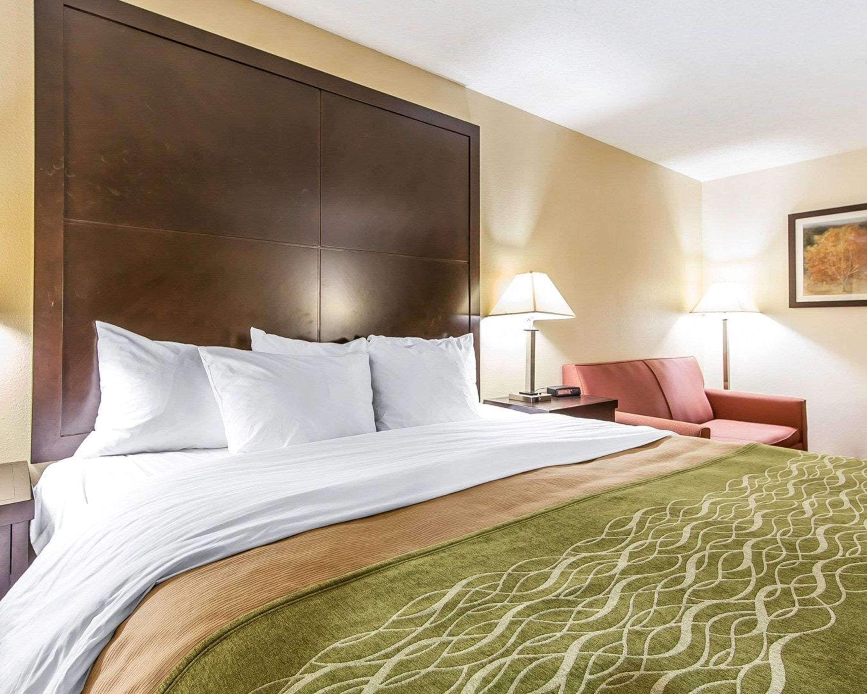 A hotel room with a large bed, a headboard, a nightstand with a lamp, a chair, and a painting on the wall.