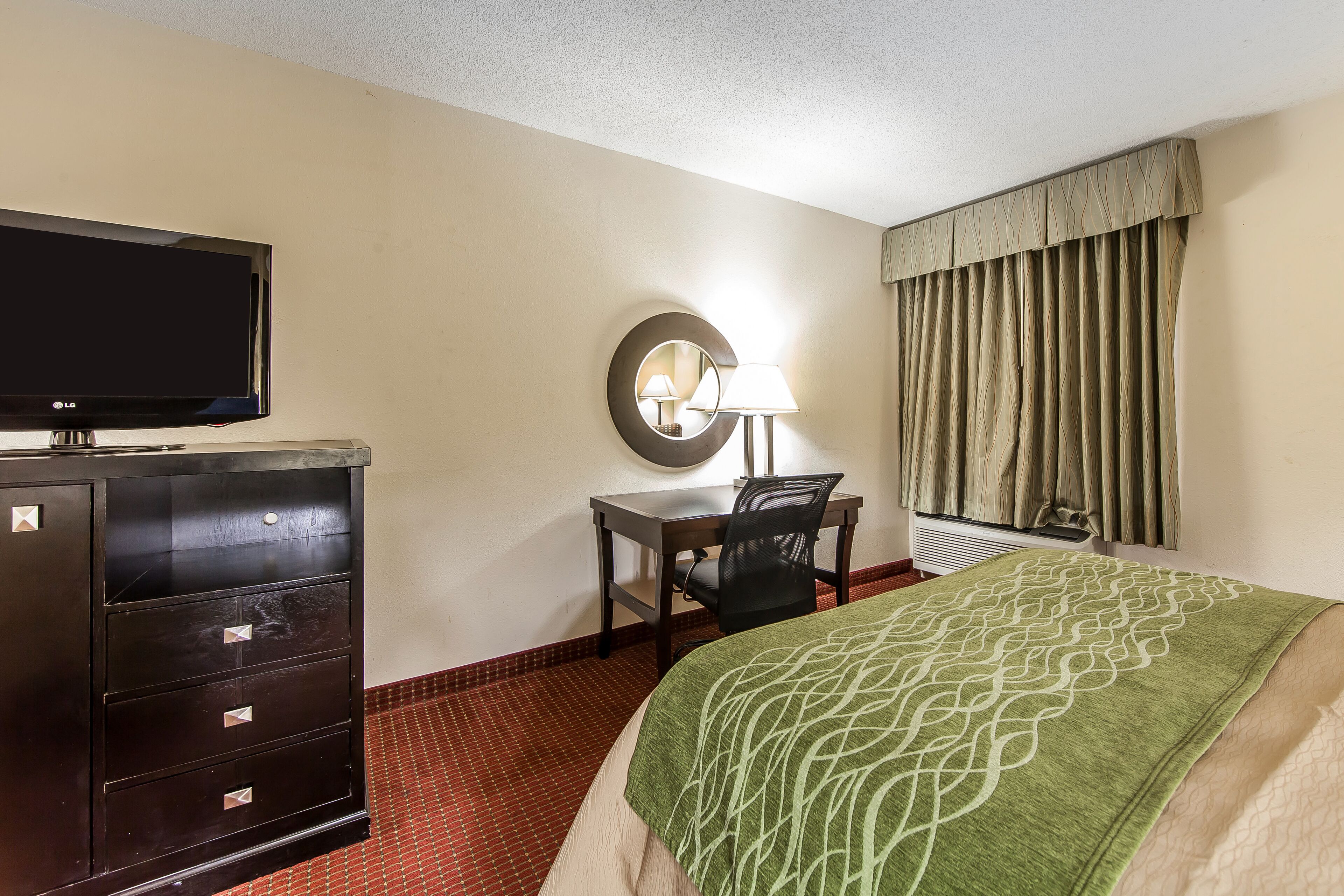 A hotel room with a bed, a desk, a television, a dresser, a lamp, and a window with curtains.