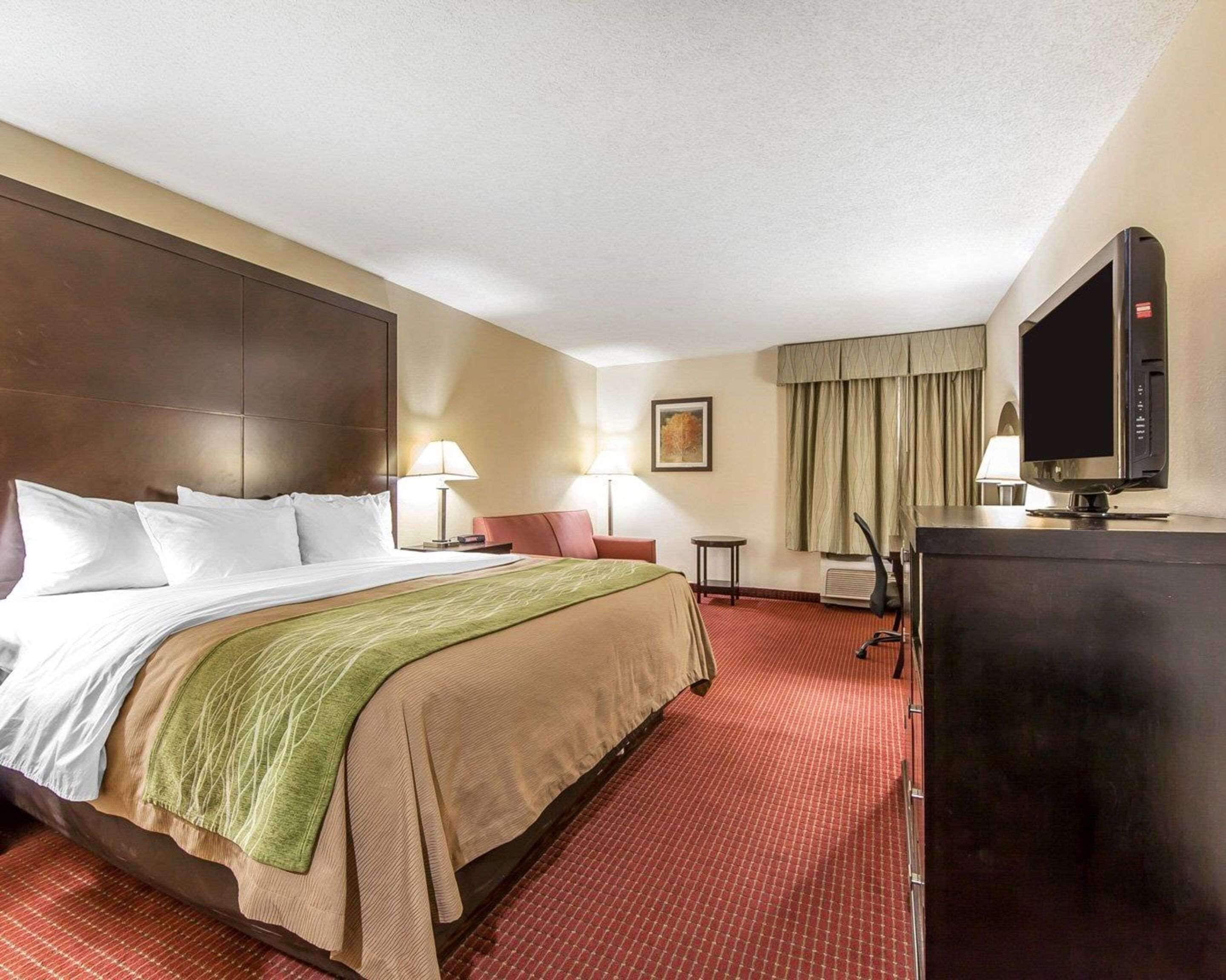 Habitación de hotel con una cama grande, un escritorio con ordenador, televisor y ventana con cortinas.