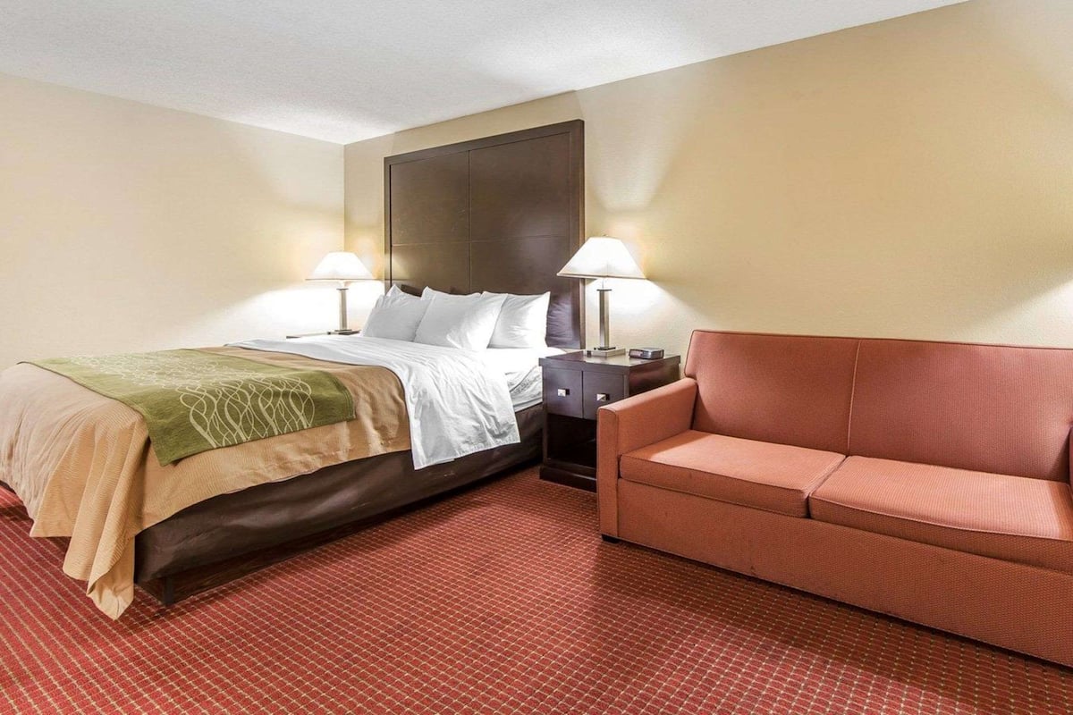 A hotel room with a bed, two bedside lamps, a nightstand, a red sofa, and a patterned carpet.