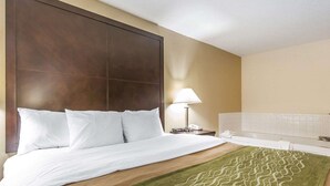 A hotel room with a large bed, a nightstand with a lamp, a bathroom with a bathtub, and a green patterned rug.