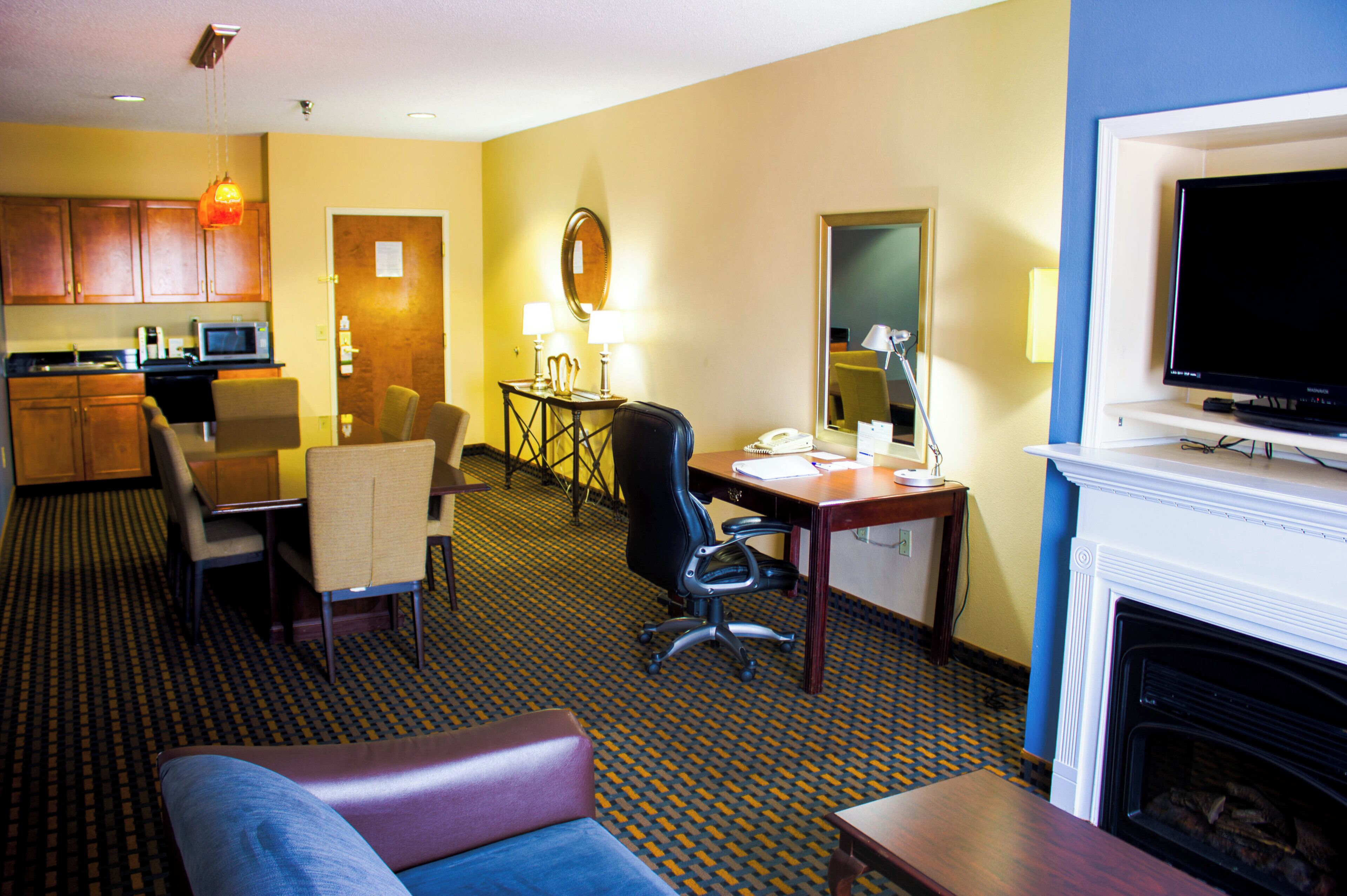 A hotel room with a sofa, a dining table with chairs, a kitchen area, a television, and a fireplace.