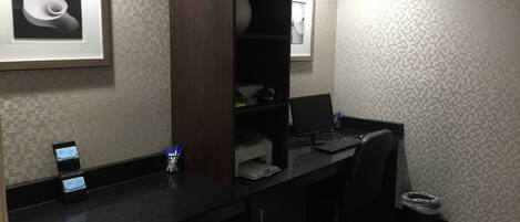 Quarto de hotel com escrivaninha, computador, cadeira e uma moldura na parede.