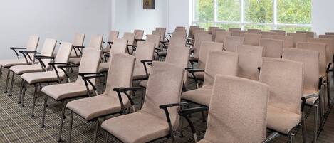 A conference room with rows of beige chairs, a carpeted floor, a large window with a view of trees, and a white wall with a framed picture.