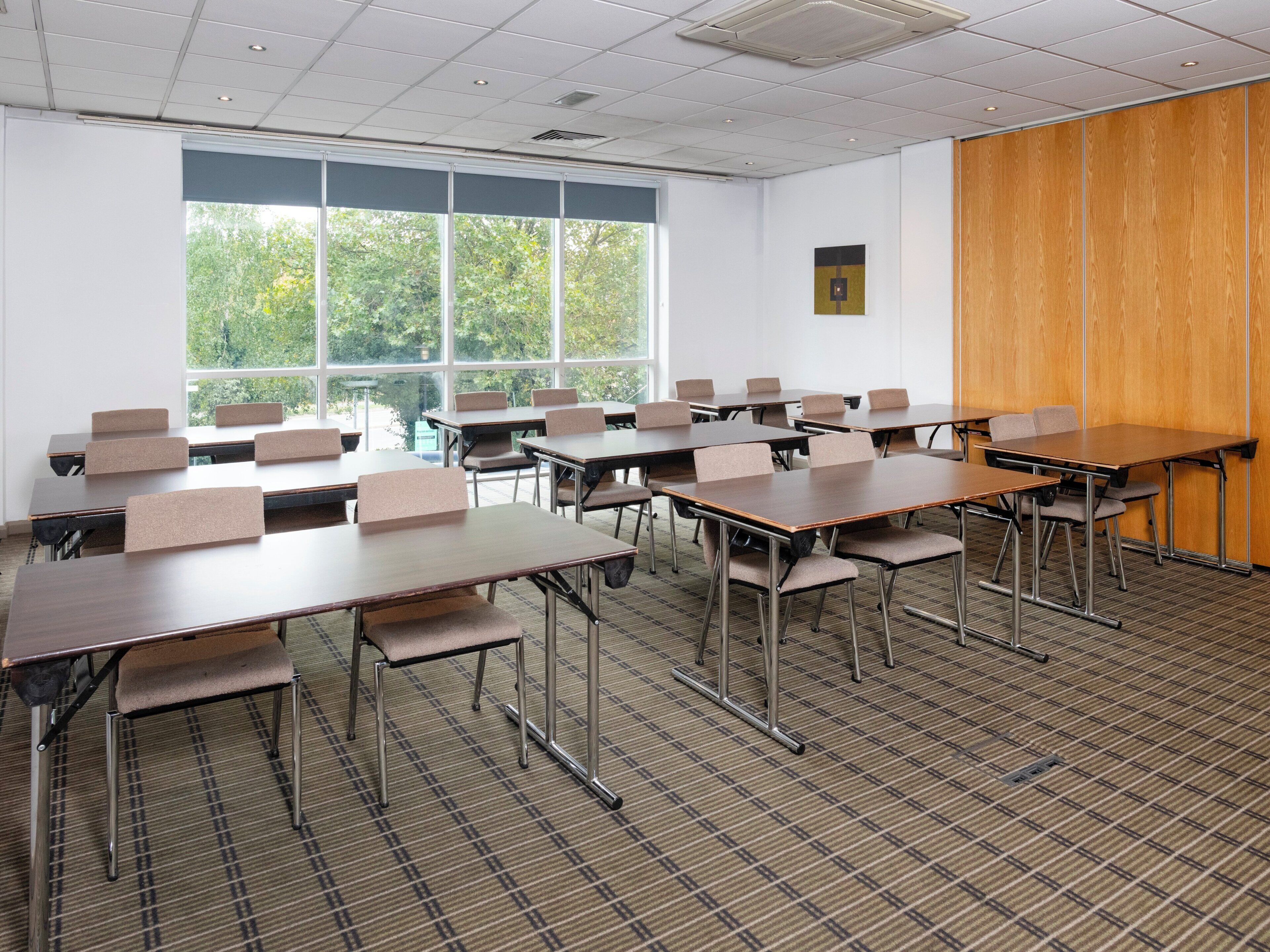 A classroom with desks and chairs arranged in rows, large windows with a view of trees, and a wooden panel on one wall.