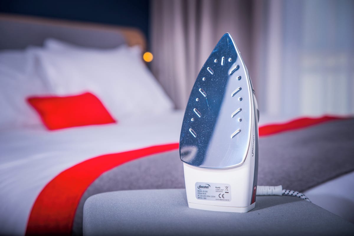 A hotel room with a bed, a red pillow, and a white iron on a bedside table.
