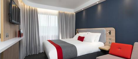 A hotel room with a bed, a TV mounted on the wall, a window with curtains, and a red and grey sofa.