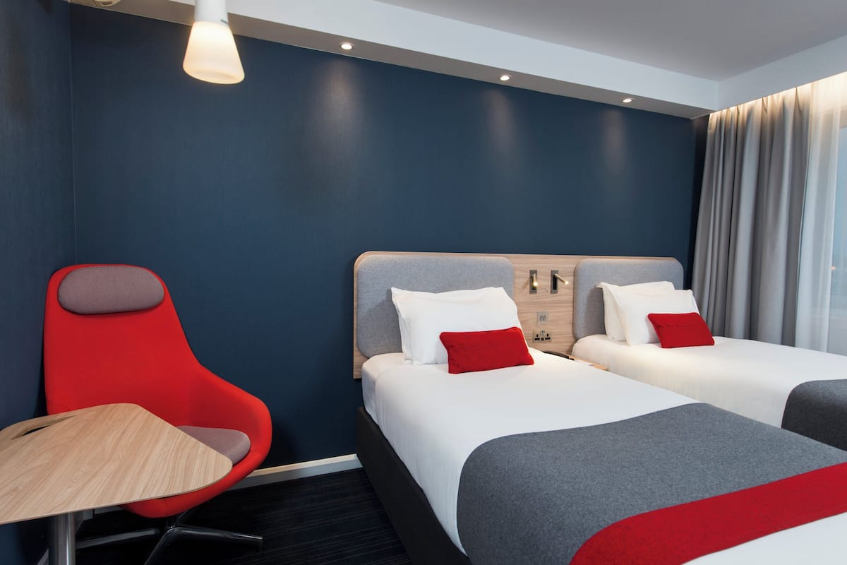 A hotel room with two beds, a red chair, a wooden table, and a modern light fixture.