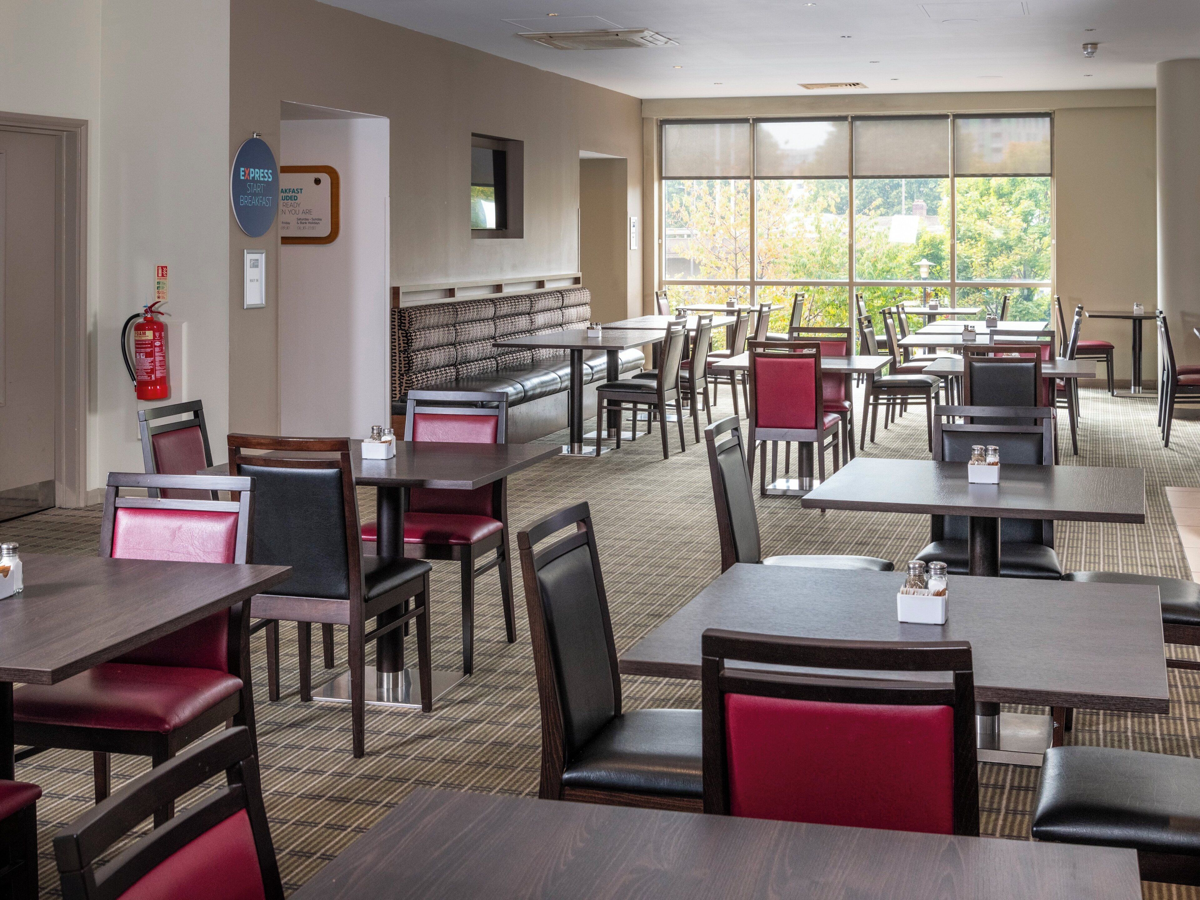 A hotel dining area with multiple tables and chairs, a fire extinguisher, and a view of trees through large windows.