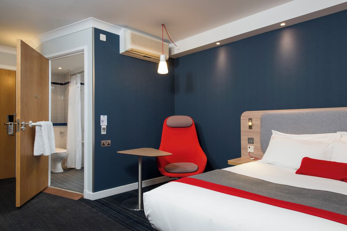 A hotel room with a bed, a red chair, a small table, a shower area, and an air conditioning unit.