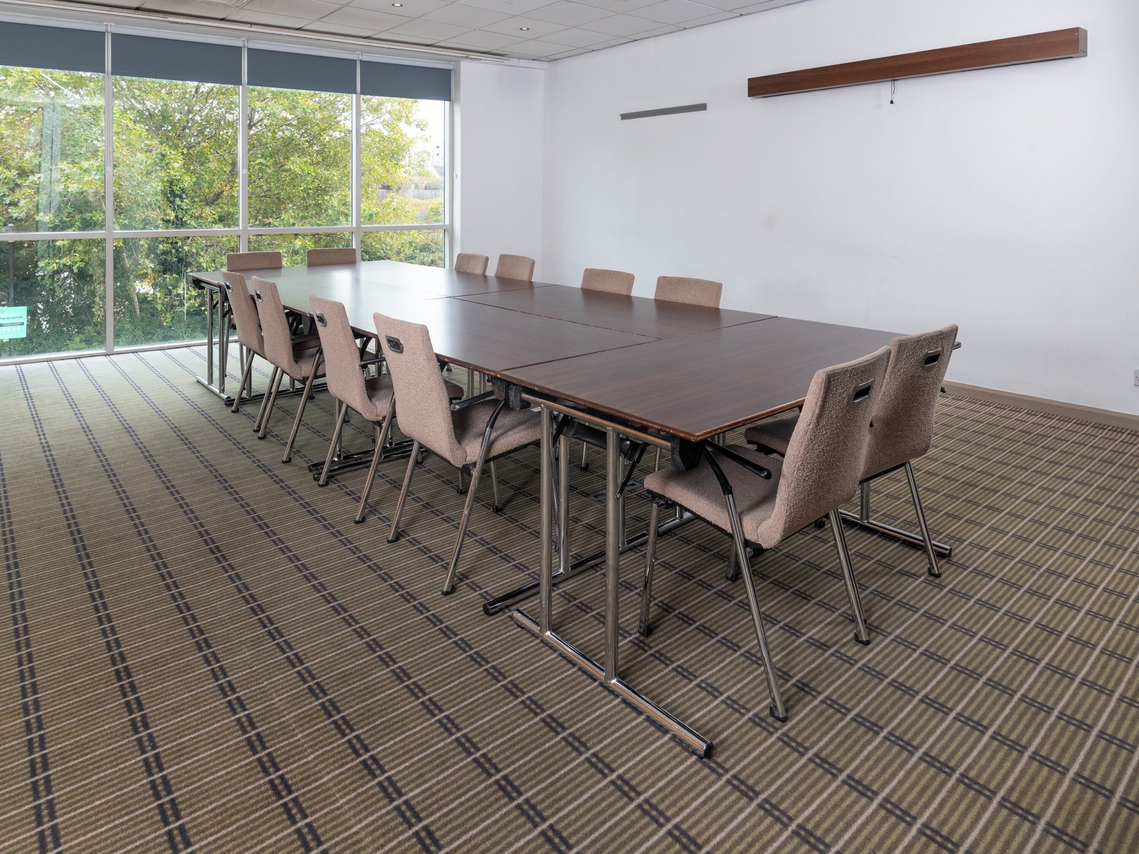 A conference room with a long table and six chairs, large windows with a view of trees, and a carpeted floor.