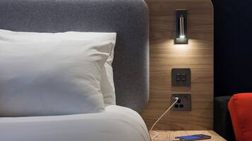Un lit bien fait, avec une tête de lit, une table de chevet dotée d’un câble de chargement et un luminaire fixé au mur.