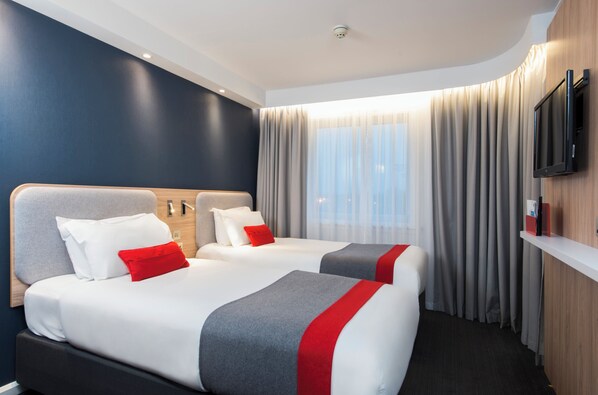 Hotellihuone, jossa on kaksi sänkyä, taulutelevisio, puinen pääty ja suuri ikkuna, jossa on verhot.