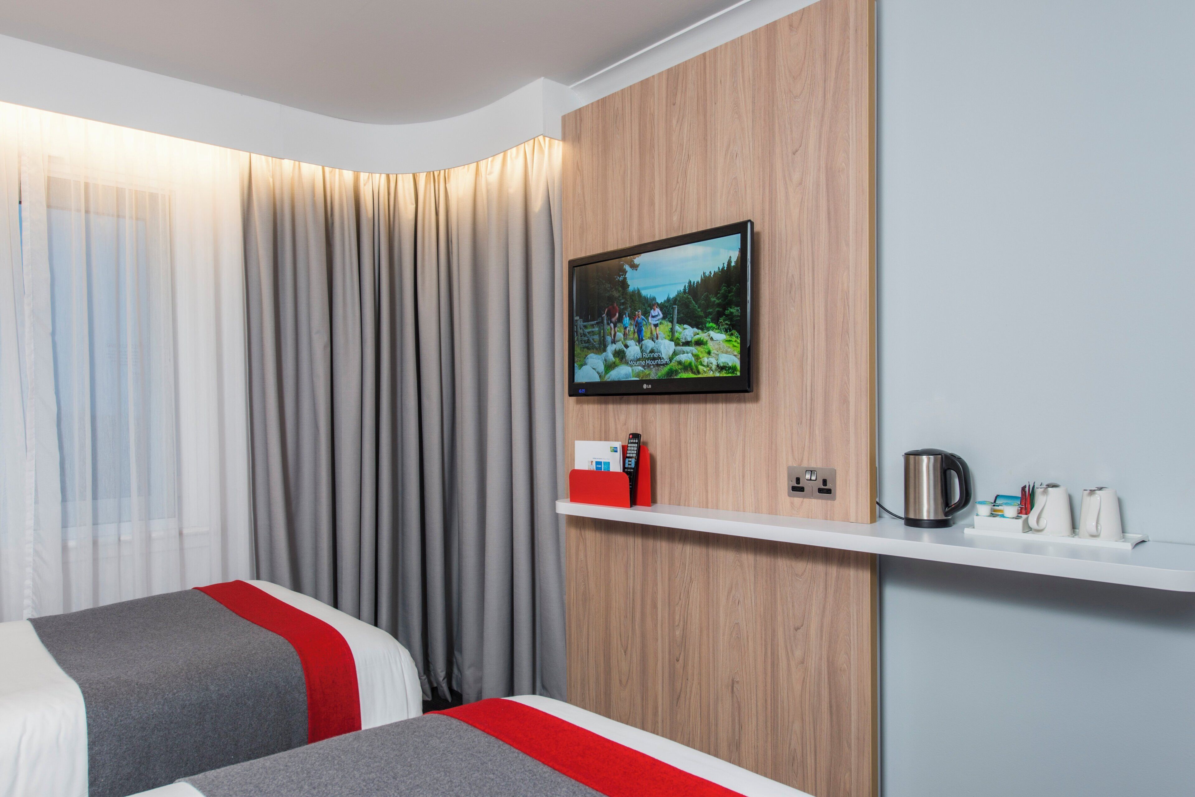 A hotel room with two beds, a television mounted on the wall, a shelf with a kettle and cups, and a window with curtains.