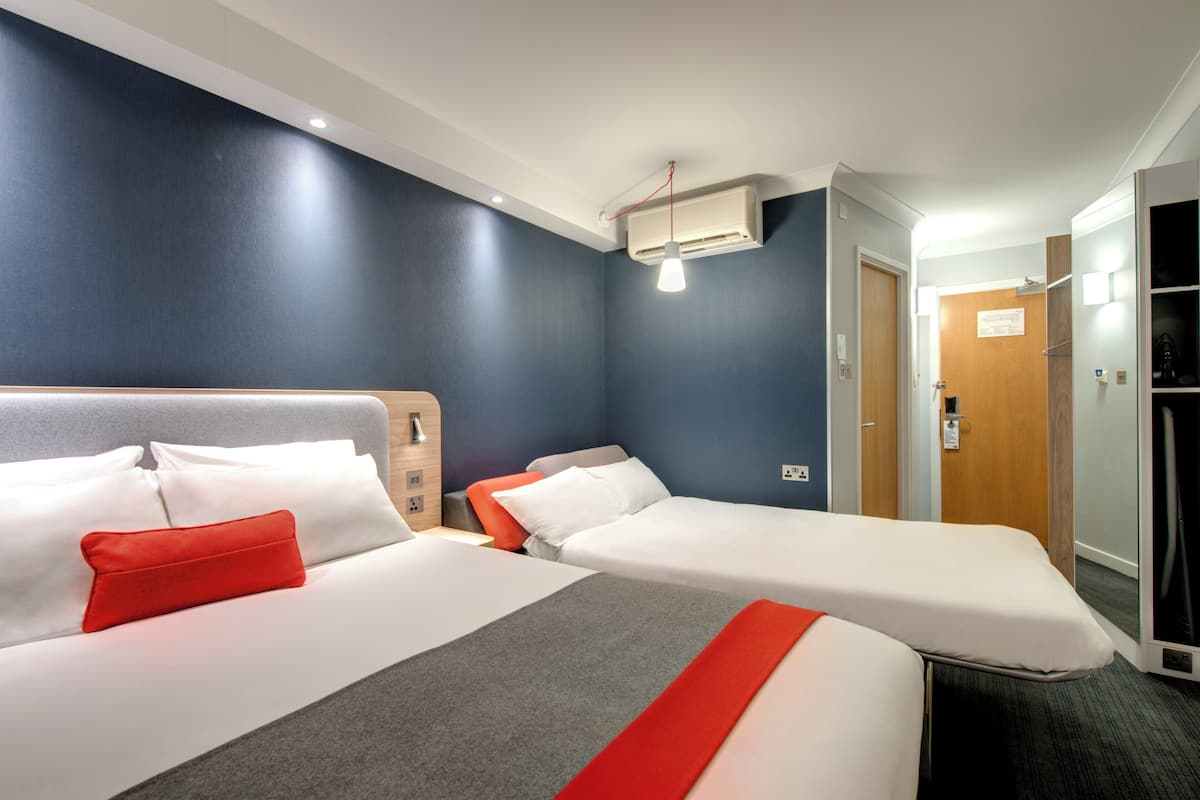 A hotel room with two beds, a red pillow, a grey blanket, and a white headboard.