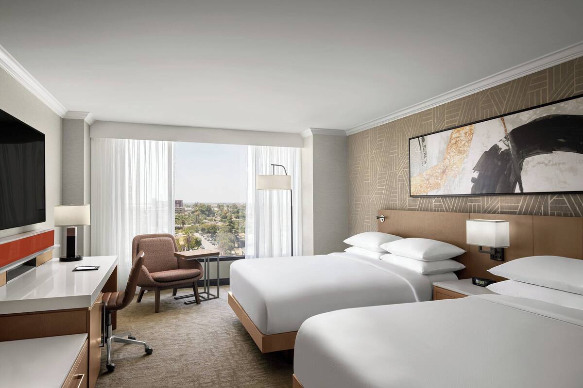 A hotel room with two beds, a desk, a chair, a TV, and a large window with curtains.