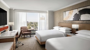 A hotel room with two beds, a desk, a chair, a TV, and a large window with curtains.