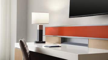 A modern hotel room with a desk, a chair, a lamp, and a flat-screen TV mounted on the wall.
